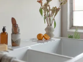 A white bathtub with dried flowers in a vase and oranges on the edge, next to soap and a brown bottle.