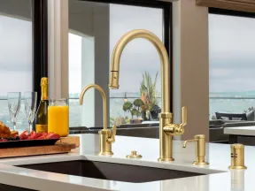 Modern kitchen sink with gold faucets and soap dispenser overlooking ocean view through large windows.