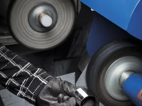 Worker wearing gloves polishing a metal ring using an industrial grinding machine with spinning wheels.