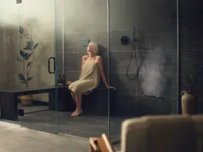 Woman wrapped in towel relaxing inside a modern glass steam shower with dark tiles and indoor plants.