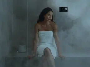 Woman in white towel relaxing on a steam room bench with a control panel on the wall amid steam.