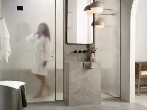 Modern bathroom with textured glass shower, concrete sink, pendant lights, and a woman in a white robe.