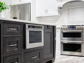 Modern kitchen with dark island, built-in stainless steel microwave, stainless steel stove, white cabinetry, and tiled backsplash.