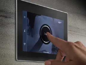 Hand pressing start button on a sleek wall-mounted touchscreen control panel with blue interface.