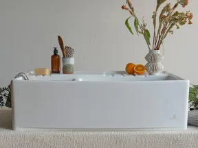 Modern white ceramic farmhouse sink on textured beige cloth with soap, brush, oranges, and vase with dried flowers.