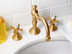 Brushed gold faucet with separate hot and cold handles on white bathroom sink with tiled wall background