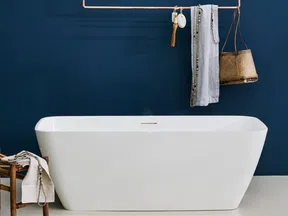 Modern white freestanding bathtub against a deep blue wall with hanging towels and a wicker basket