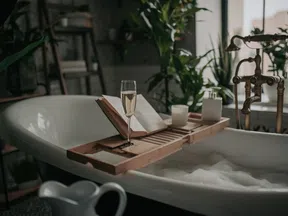 Bathtub filled with bubbles, wooden tray holding a book, candle, and glass of champagne surrounded by plants.