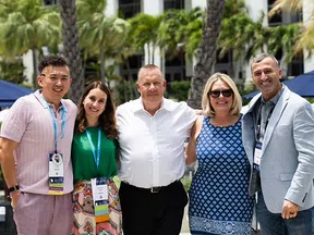 Group of five smiling adults standing outdoors with palm trees and umbrellas in the background at a sunny event.