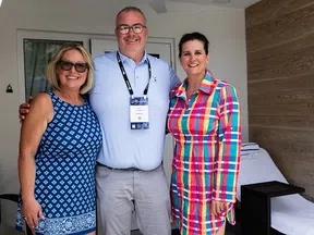 Three smiling adults posing indoors, one in a blue patterned dress, one in a white shirt and badge, and one in a colorful plaid dress.