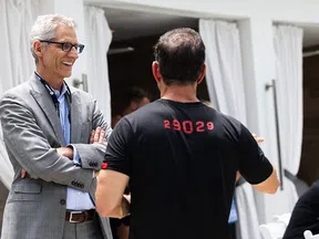 Two men engaged in conversation, one wearing a gray suit and glasses, the other in a black shirt with red numbers.