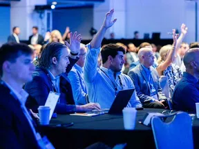 Audience in a conference room raising hands, engaging in a professional event with laptops and notebooks.