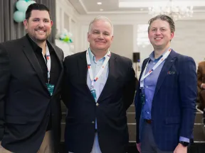 Three men in business attire smiling at a conference with balloons and stage in the background.