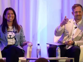 Two panelists seated on stage engaged in a discussion with microphones and water bottles in front of blue curtains.