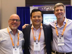 Three men wearing conference badges and orange lanyards posing together at a business event booth.