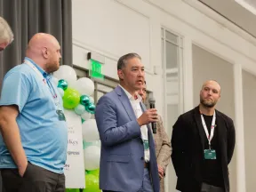 Group of five men in business attire speaking at an indoor event with balloons and signage in the background