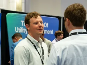 Two men wearing lanyards engaged in conversation at a professional networking event with exhibition booths.