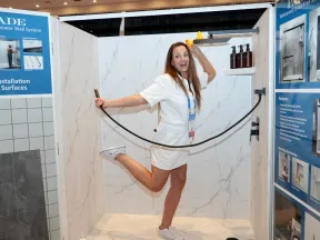 Woman demonstrating Palisade waterproof grout-free shower wall system in a marble-style shower booth at a trade show.