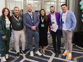 Group of six professionals posing at a conference venue with badges and casual business attire