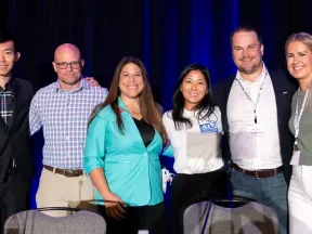 Group of six professionals smiling and posing together against a dark blue curtain backdrop at a conference.