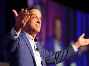 Man in blue suit speaking on stage with arms outstretched and purple-blue light background