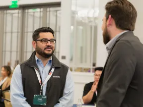 Two men engaged in a professional conversation at a networking event with attendees in the background.