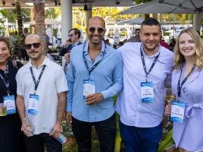 Five professionals wearing conference badges smiling outdoors at a networking event with drinks in hand