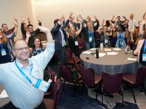 Group of conference attendees smiling and raising hands around tables in a meeting room with lanyards and badges.