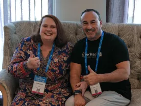 Two smiling conference attendees sitting on a sofa showing thumbs up with event badges and blue lanyards
