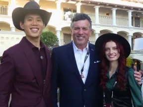 Three people smiling outdoors at a conference or event, two wearing black hats and one man in a suit.