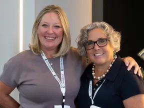 Two smiling women wearing conference badges posing together at an indoor event with a lamp in the background