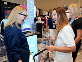 Three women engaging happily at a busy conference with booths and promotional displays in the background