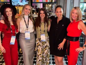 Five women at a social event posing together on black and white tiled floor, dressed in colorful and stylish outfits.