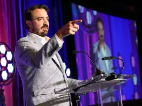 Man in gray suit speaking and pointing on stage with purple curtains and large screen backdrop.