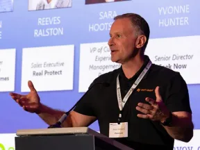 Man speaking at a conference podium with a presentation slide of speaker names and titles in the background