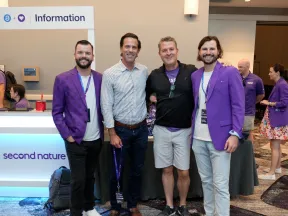 Four men smiling at an event, with two wearing purple jackets in front of an information desk for AppFolio and Second Nature.