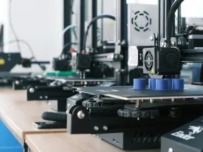 Row of 3D printers producing blue plastic prototypes on wooden tables in a bright workshop environment