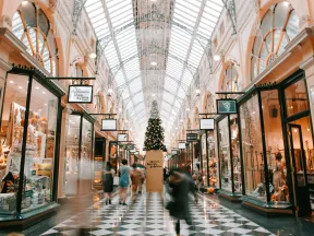 Indoor shopping arcade with Christmas decorations, high glass ceiling, and shoppers walking on checkered floor.