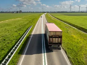 Truck driving on a rural highway surrounded by green fields and wind turbines under a blue sky with clouds.