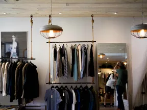 Modern boutique interior with hanging clothes racks, pendant lights, and customers shopping in front of a large mirror.