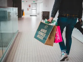 Person walking in mall hallway carrying shopping bags labeled Good Things Inside and PINK with a camera in hand.