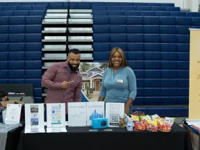 Two smiling representatives stand behind a business association table with brochures and snacks at an indoor event.