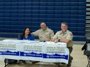 Three smiling adults, two in scout uniforms, sitting behind a Join a Troop Boy Scouts recruitment table in a gym.