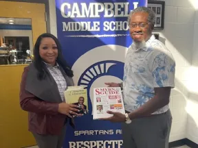 Two adults holding books in front of a Campbell Middle School banner highlighting respect and responsibility values.