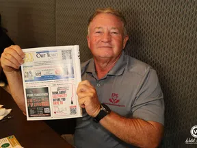 Smiling man in gray shirt holding a newspaper with various ads and articles seated at a table indoors.