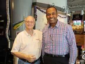 Two men smiling and posing inside a decorated bar or restaurant with festive string lights and TV screens.