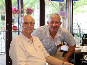 Two elderly men smiling indoors near windows with hanging flowers and tables in the background