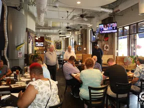 People dining and socializing in a bright, casual restaurant interior with wooden decor and hanging lights.
