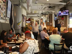 Diverse group of people seated and two men standing in a busy restaurant with wooden decor and TV screens.