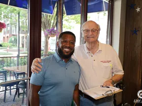 Two men smiling together indoors, one holding a clipboard, with large windows and outdoor seating visible.
